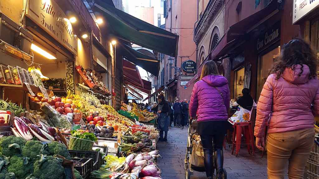 Italian street market with fresh produce stalls and shoppers walking through a historic alley